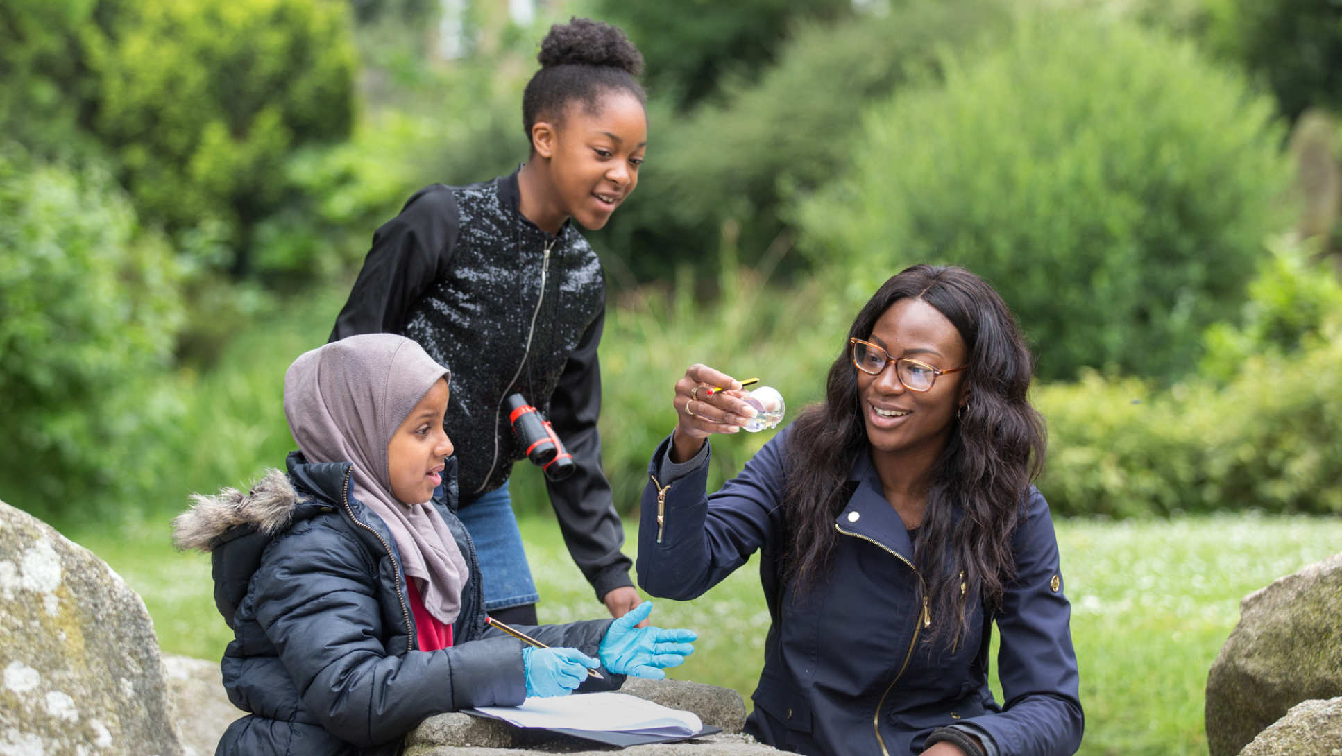 Two children and a woman recording details of the insects they have found in a park.