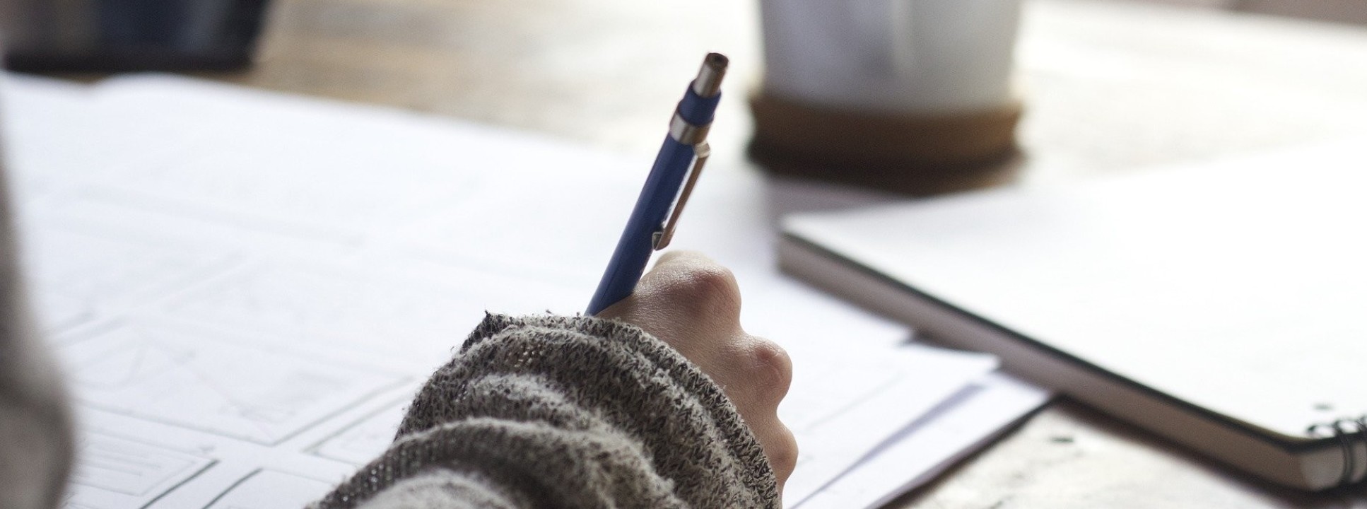 Close-up photograph of a person writing at a desk with a mug in the background