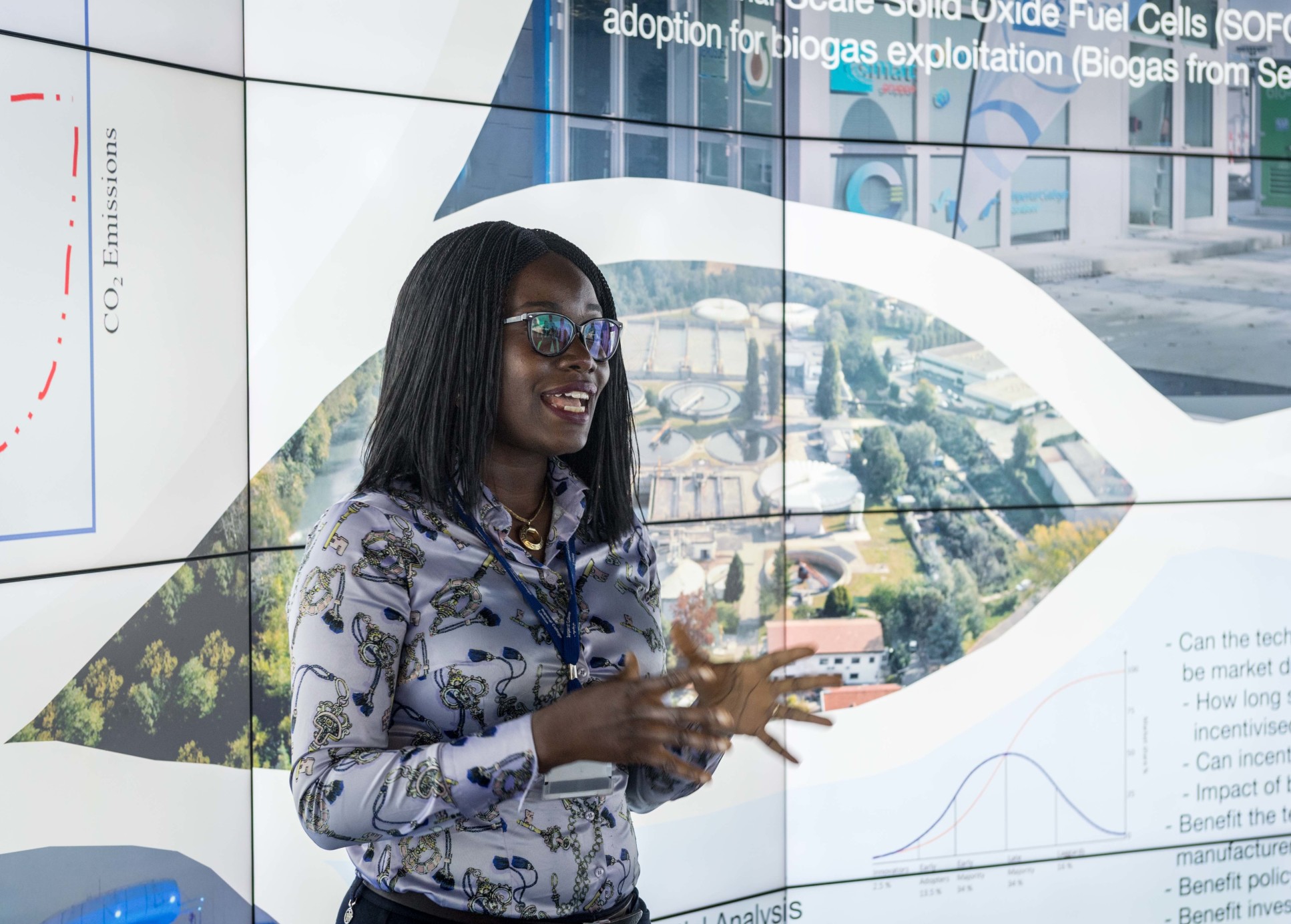 Gbemi Oluleye, a black woman in her mid thirties, addresses an audience, she stands in front of a video screen showing an aerial photo of an industrial plant. Gbemi wears a light blouse and has dark thinly braided hair and glasses.