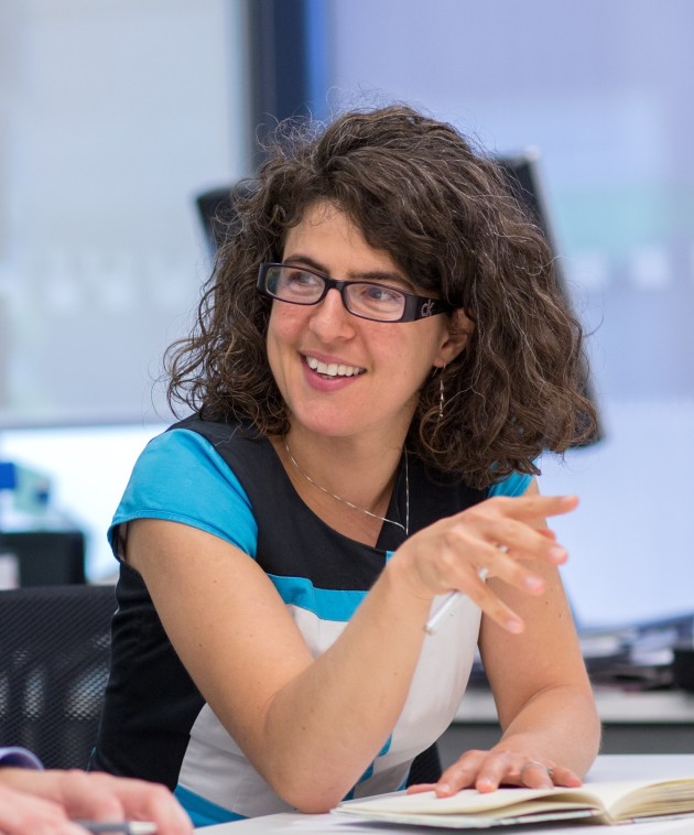 Alyssa Gilbert, a white woman in her early forties, talks to a room of people. She has dark curly hair, glasses and wears a blue and white dress